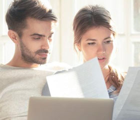 Pareja sentada en el sofá mientras revisan diferentes documentos físicos y en el ordenador